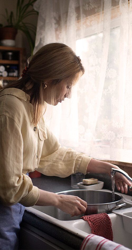 A woman fills a pot with water at kitchen sink near a window.