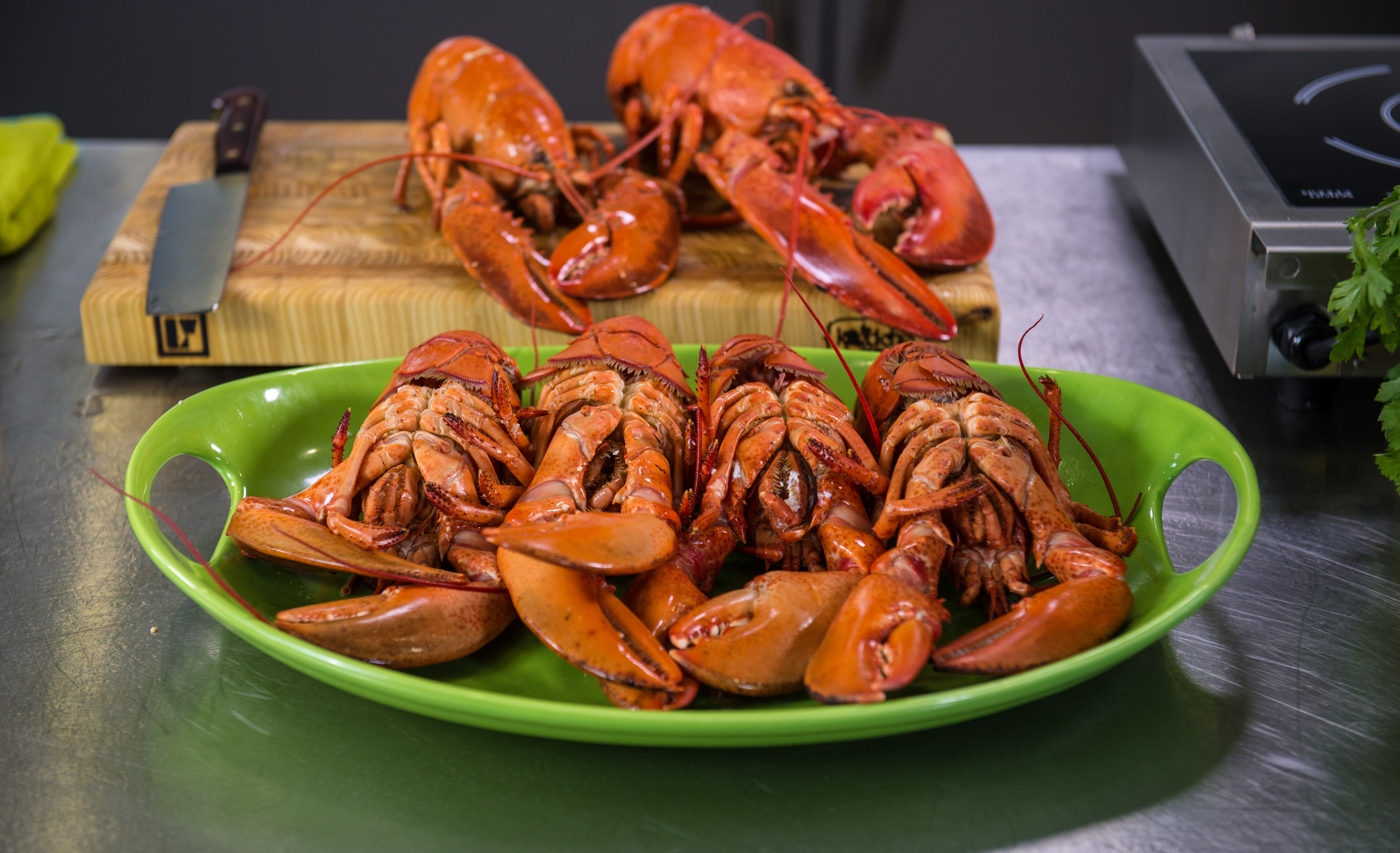 A photo of a green plate with multiple whole cooked lobsters, as well as some lobsters placed on a cutting board behind it.